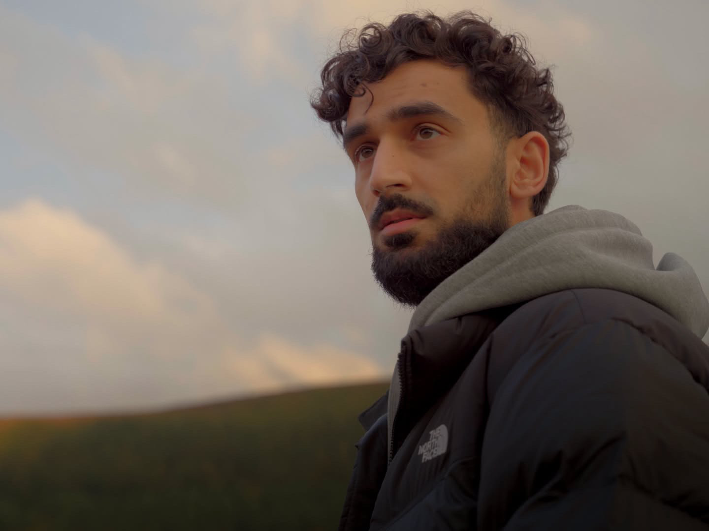 Close-up portrait of filmmaker Hamza Shaf, curly hair and beard, looking off to the left against a soft evening sky