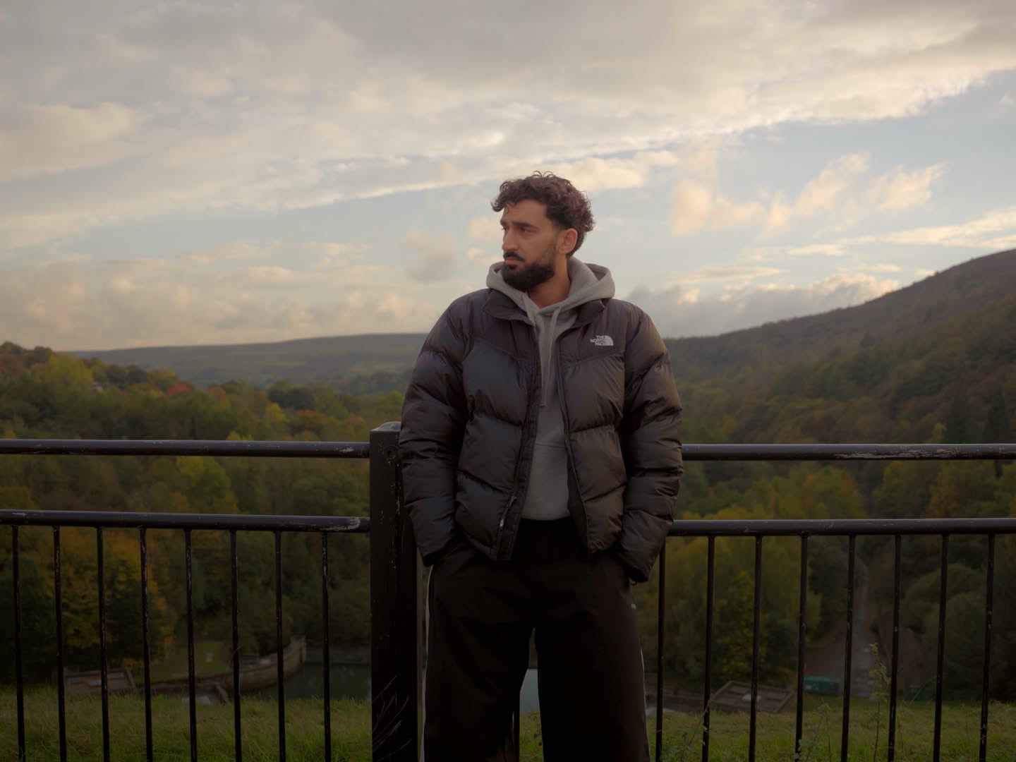 Hamza Shaf standing at a railing above an autumn valley of green and amber woodland under a cloudy sky